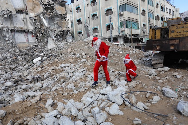   فلسطيني وابنه يرتديان زي بابا نويل يسيران بالقرب من ركام برج الجوهرة الذي تعرض للقصف خلال التصعيد الإسرائيلي الأخير على قطاع غزة.