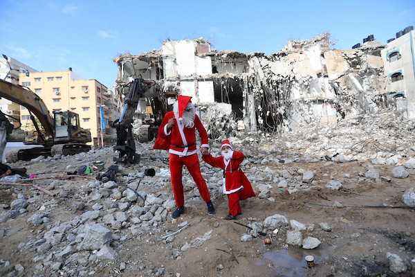   فلسطيني وابنه يرتديان زي بابا نويل يسيران بالقرب من ركام برج الجوهرة الذي تعرض للقصف خلال التصعيد الإسرائيلي الأخير على قطاع غزة.