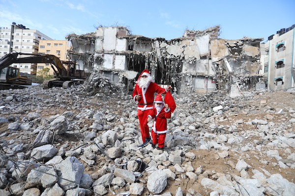   فلسطيني وابنه يرتديان زي بابا نويل يسيران بالقرب من ركام برج الجوهرة الذي تعرض للقصف خلال التصعيد الإسرائيلي الأخير على قطاع غزة.