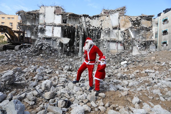   فلسطيني وابنه يرتديان زي بابا نويل يسيران بالقرب من ركام برج الجوهرة الذي تعرض للقصف خلال التصعيد الإسرائيلي الأخير على قطاع غزة.