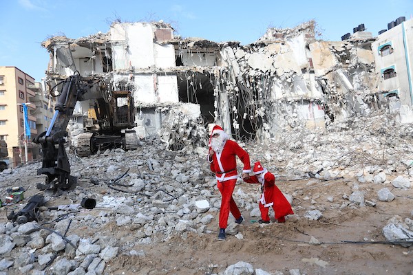   فلسطيني وابنه يرتديان زي بابا نويل يسيران بالقرب من ركام برج الجوهرة الذي تعرض للقصف خلال التصعيد الإسرائيلي الأخير على قطاع غزة.