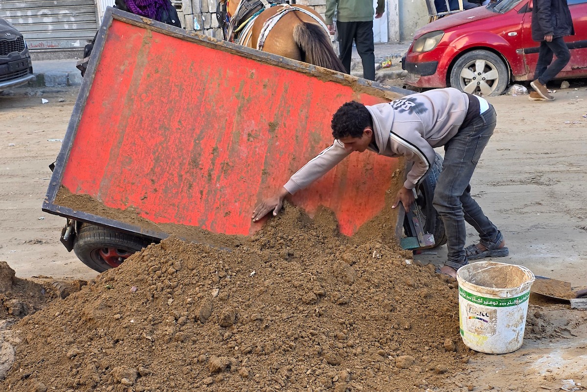 في خان يونس، جنوب قطاع غزة، يقوم أصحاب المتاجر الفلسطينيون بإزالة الأنقاض بأيديهم، ويعيدون بناء متاجرهم المتضررة باستخدام الحجارة والطين الأحمر، 27 ديسمبر/كانون الأول 2025 (صورة: طارق محمد)