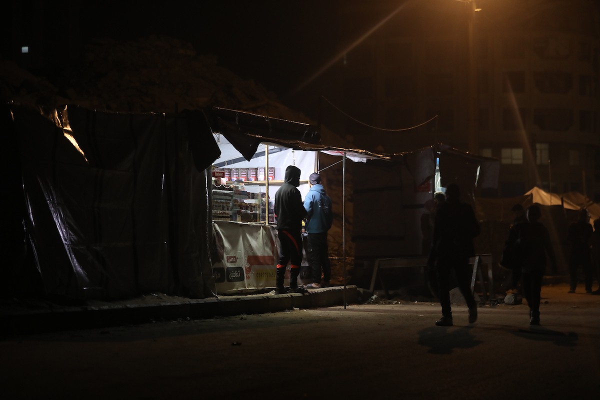 يسير فلسطينيون في أحد شوارع مدينة غزة، بينما يكافح النازحون في خيامهم لمواجهة الظلام، يتدفؤون ويطهون طعامهم بالحطب، محرومين حتى من أبسط مقومات الحياة، 23 يناير/كانون الثاني 2020. صورة: عمر أشتوي