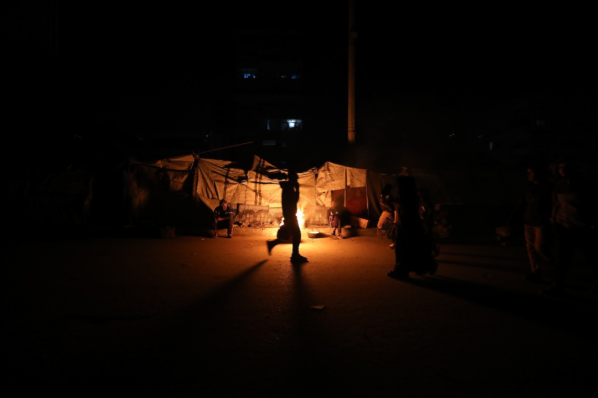 يسير فلسطينيون في أحد شوارع مدينة غزة، بينما يكافح النازحون في خيامهم لمواجهة الظلام، يتدفؤون ويطهون طعامهم بالحطب، محرومين حتى من أبسط مقومات الحياة، 23 يناير/كانون الثاني 2020. صورة: عمر أشتوي