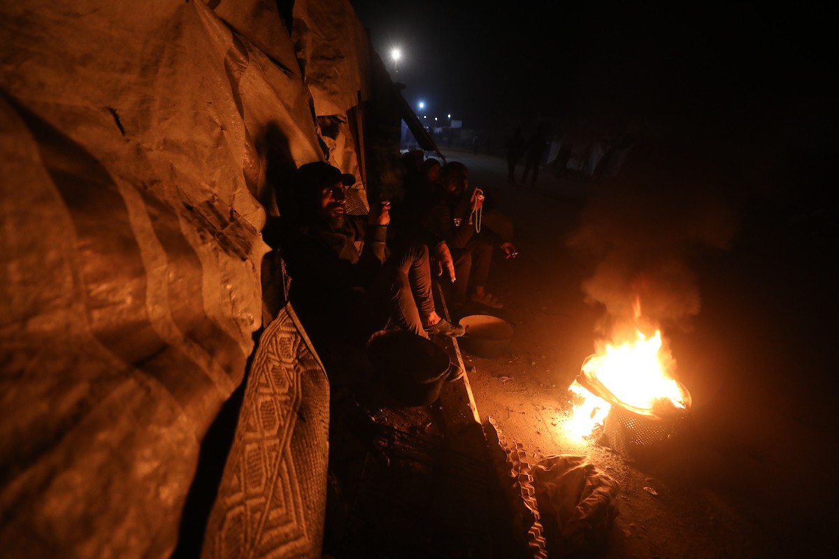 يسير فلسطينيون في أحد شوارع مدينة غزة، بينما يكافح النازحون في خيامهم لمواجهة الظلام، يتدفؤون ويطهون طعامهم بالحطب، محرومين حتى من أبسط مقومات الحياة، 23 يناير/كانون الثاني 2020. صورة: عمر أشتوي