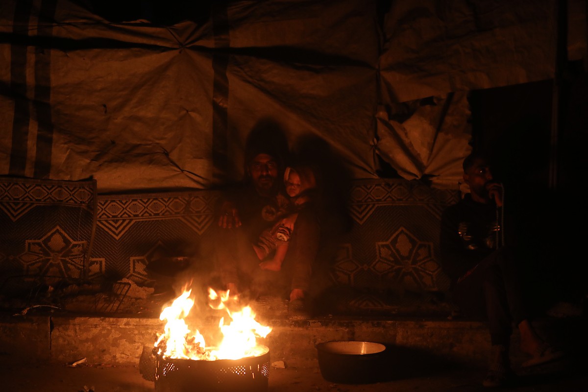 يسير فلسطينيون في أحد شوارع مدينة غزة، بينما يكافح النازحون في خيامهم لمواجهة الظلام، يتدفؤون ويطهون طعامهم بالحطب، محرومين حتى من أبسط مقومات الحياة، 23 يناير/كانون الثاني 2020. صورة: عمر أشتوي