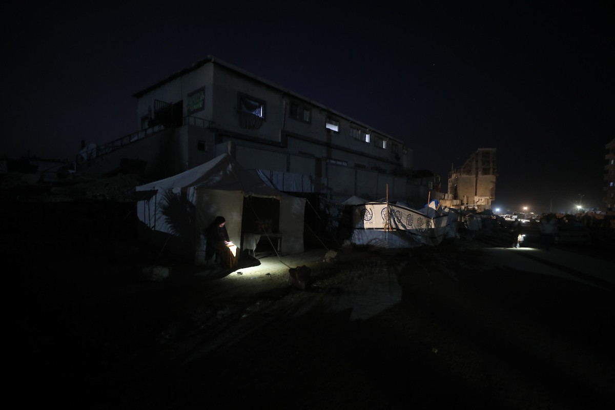 يسير فلسطينيون في أحد شوارع مدينة غزة، بينما يكافح النازحون في خيامهم لمواجهة الظلام، يتدفؤون ويطهون طعامهم بالحطب، محرومين حتى من أبسط مقومات الحياة، 23 يناير/كانون الثاني 2020. صورة: عمر أشتوي