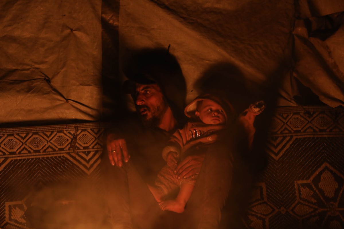 يسير فلسطينيون في أحد شوارع مدينة غزة، بينما يكافح النازحون في خيامهم لمواجهة الظلام، يتدفؤون ويطهون طعامهم بالحطب، محرومين حتى من أبسط مقومات الحياة، 23 يناير/كانون الثاني 2020. صورة: عمر أشتوي