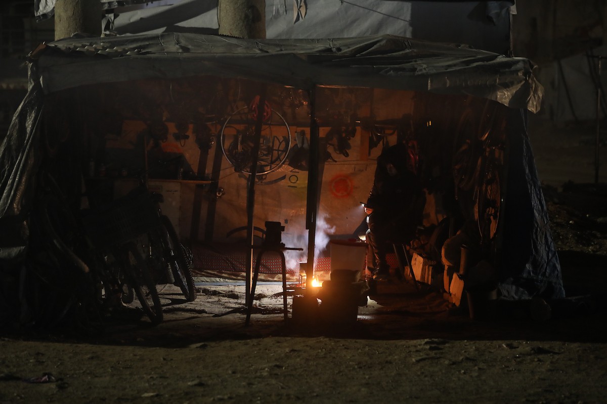 يسير فلسطينيون في أحد شوارع مدينة غزة، بينما يكافح النازحون في خيامهم لمواجهة الظلام، يتدفؤون ويطهون طعامهم بالحطب، محرومين حتى من أبسط مقومات الحياة، 23 يناير/كانون الثاني 2020. صورة: عمر أشتوي