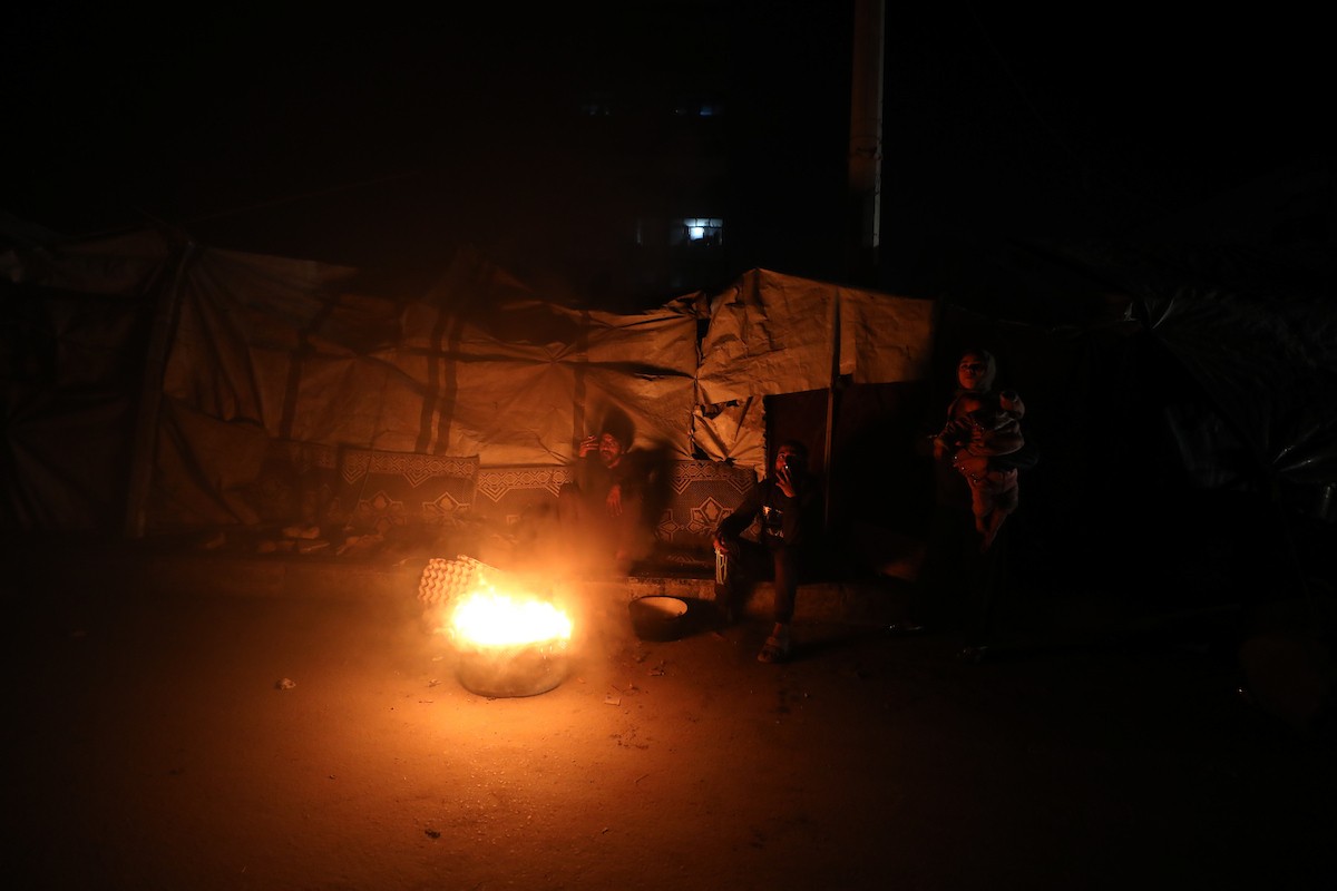 يسير فلسطينيون في أحد شوارع مدينة غزة، بينما يكافح النازحون في خيامهم لمواجهة الظلام، يتدفؤون ويطهون طعامهم بالحطب، محرومين حتى من أبسط مقومات الحياة، 23 يناير/كانون الثاني 2020. صورة: عمر أشتوي