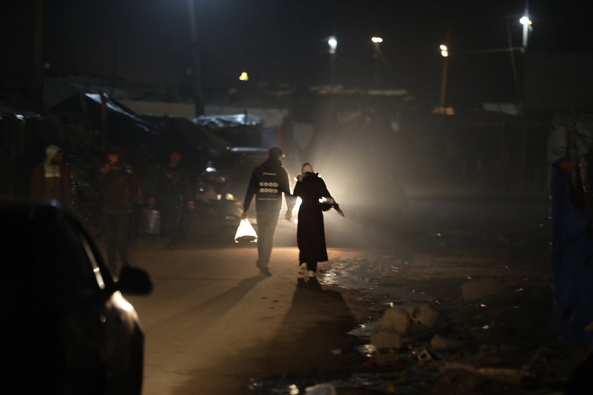 يسير فلسطينيون في أحد شوارع مدينة غزة، بينما يكافح النازحون في خيامهم لمواجهة الظلام، يتدفؤون ويطهون طعامهم بالحطب، محرومين حتى من أبسط مقومات الحياة، 23 يناير/كانون الثاني 2020. صورة: عمر أشتوي