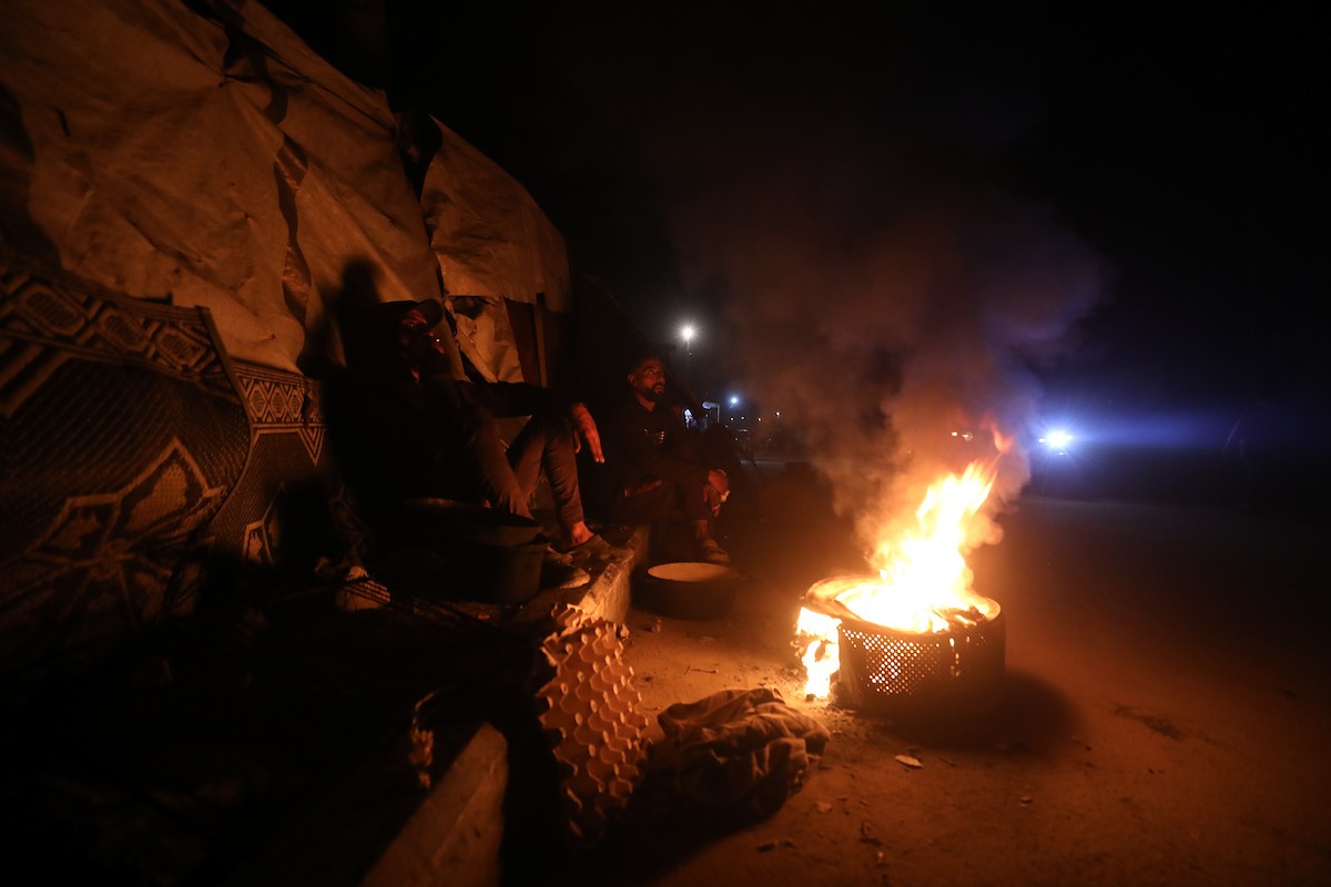 يسير فلسطينيون في أحد شوارع مدينة غزة، بينما يكافح النازحون في خيامهم لمواجهة الظلام، يتدفؤون ويطهون طعامهم بالحطب، محرومين حتى من أبسط مقومات الحياة، 23 يناير/كانون الثاني 2020. صورة: عمر أشتوي