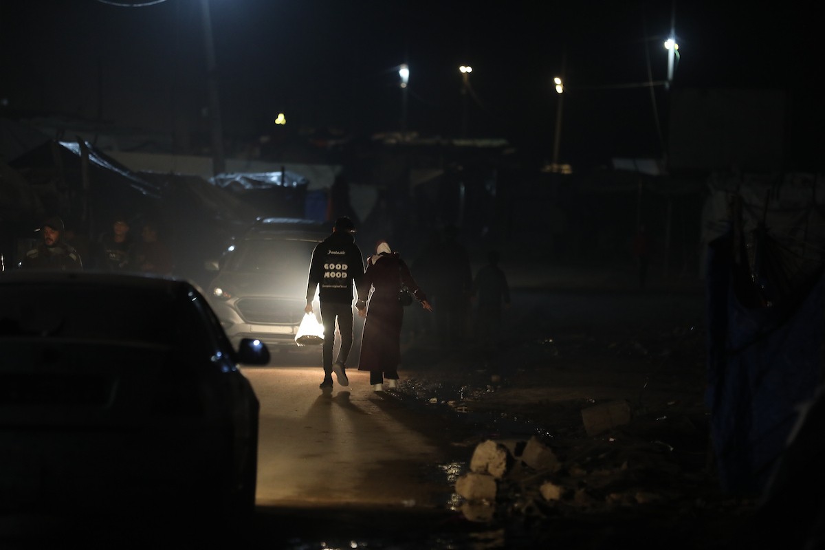 يسير فلسطينيون في أحد شوارع مدينة غزة، بينما يكافح النازحون في خيامهم لمواجهة الظلام، يتدفؤون ويطهون طعامهم بالحطب، محرومين حتى من أبسط مقومات الحياة، 23 يناير/كانون الثاني 2020. صورة: عمر أشتوي