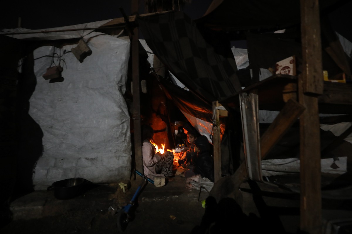 يسير فلسطينيون في أحد شوارع مدينة غزة، بينما يكافح النازحون في خيامهم لمواجهة الظلام، يتدفؤون ويطهون طعامهم بالحطب، محرومين حتى من أبسط مقومات الحياة، 23 يناير/كانون الثاني 2020. صورة: عمر أشتوي