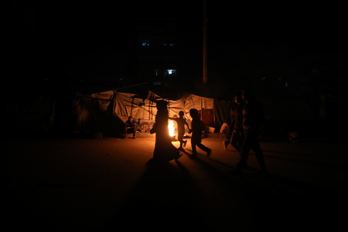 يسير فلسطينيون في أحد شوارع مدينة غزة، بينما يكافح النازحون في خيامهم لمواجهة الظلام، يتدفؤون ويطهون طعامهم بالحطب، محرومين حتى من أبسط مقومات الحياة، 23 يناير/كانون الثاني 2020. صورة: عمر أشتوي