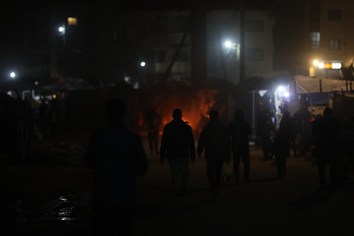 يسير فلسطينيون في أحد شوارع مدينة غزة، بينما يكافح النازحون في خيامهم لمواجهة الظلام، يتدفؤون ويطهون طعامهم بالحطب، محرومين حتى من أبسط مقومات الحياة، 23 يناير/كانون الثاني 2020. صورة: عمر أشتوي