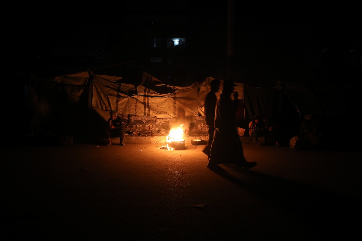 يسير فلسطينيون في أحد شوارع مدينة غزة، بينما يكافح النازحون في خيامهم لمواجهة الظلام، يتدفؤون ويطهون طعامهم بالحطب، محرومين حتى من أبسط مقومات الحياة، 23 يناير/كانون الثاني 2020. صورة: عمر أشتوي