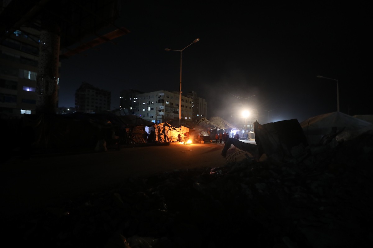 يسير فلسطينيون في أحد شوارع مدينة غزة، بينما يكافح النازحون في خيامهم لمواجهة الظلام، يتدفؤون ويطهون طعامهم بالحطب، محرومين حتى من أبسط مقومات الحياة، 23 يناير/كانون الثاني 2020. صورة: عمر أشتوي