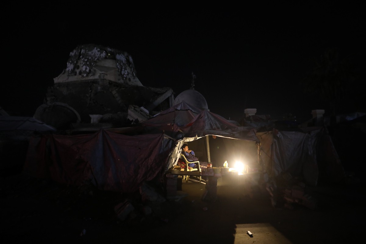 يسير فلسطينيون في أحد شوارع مدينة غزة، بينما يكافح النازحون في خيامهم لمواجهة الظلام، يتدفؤون ويطهون طعامهم بالحطب، محرومين حتى من أبسط مقومات الحياة، 23 يناير/كانون الثاني 2020. صورة: عمر أشتوي