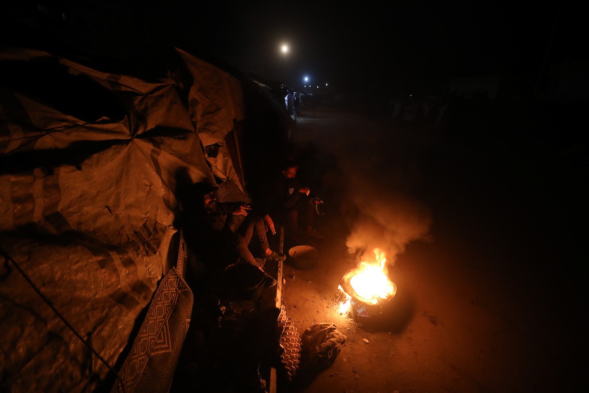 يسير فلسطينيون في أحد شوارع مدينة غزة، بينما يكافح النازحون في خيامهم لمواجهة الظلام، يتدفؤون ويطهون طعامهم بالحطب، محرومين حتى من أبسط مقومات الحياة، 23 يناير/كانون الثاني 2020. صورة: عمر أشتوي