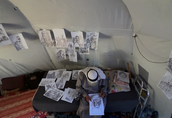 محمد زملط، رجل فلسطيني مسنّ اقتلعته الحرب من مخيم جباليا في شمال قطاع غزة، يحمل اليوم ريشته كوسيلة للبقاء، لا للفن فقط. في خيمته المتواضعة بمخيم البريج وسط القطاع، 12 أبريل/نيسان 2026، عدسة: يوسف أبو وطفة