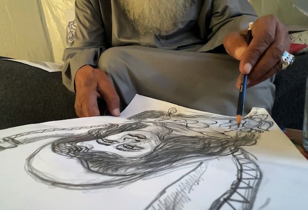 محمد زملط، رجل فلسطيني مسنّ اقتلعته الحرب من مخيم جباليا في شمال قطاع غزة، يحمل اليوم ريشته كوسيلة للبقاء، لا للفن فقط. في خيمته المتواضعة بمخيم البريج وسط القطاع، 12 أبريل/نيسان 2026، عدسة: يوسف أبو وطفة