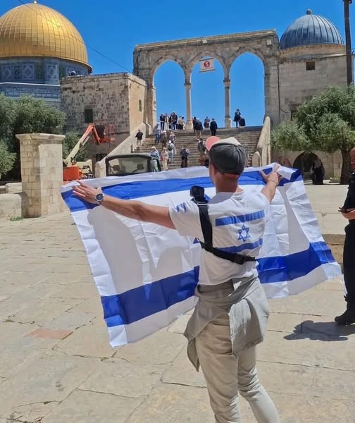 مستعمر يرتدي قميصاً يحمل  العلم الإسرائيلي ويُقدم على رفع العلم أمام قبة الصخرة المشرفة عند الرواق الغربي للمسجد الأقصى المبارك.jpg