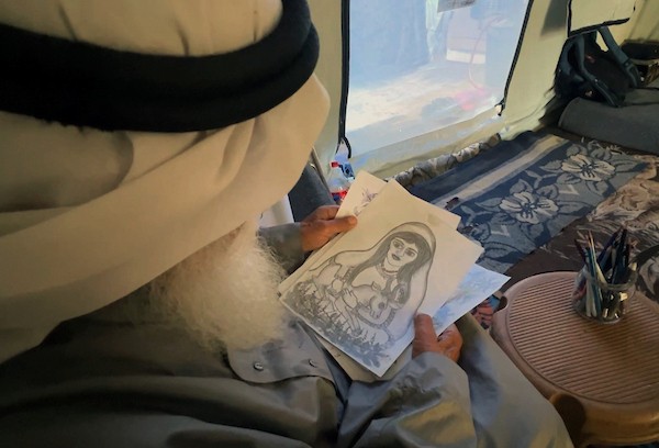 محمد زملط، رجل فلسطيني مسنّ اقتلعته الحرب من مخيم جباليا في شمال قطاع غزة، يحمل اليوم ريشته كوسيلة للبقاء، لا للفن فقط. في خيمته المتواضعة بمخيم البريج وسط القطاع، 12 أبريل/نيسان 2026، عدسة: يوسف أبو وطفة