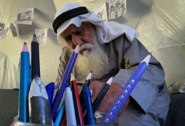 محمد زملط، رجل فلسطيني مسنّ اقتلعته الحرب من مخيم جباليا في شمال قطاع غزة، يحمل اليوم ريشته كوسيلة للبقاء، لا للفن فقط. في خيمته المتواضعة بمخيم البريج وسط القطاع، 12 أبريل/نيسان 2026، عدسة: يوسف أبو وطفة