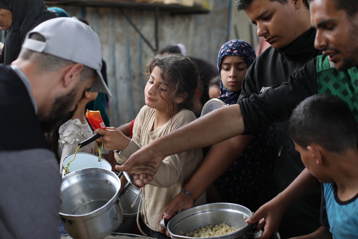 فلسطينيون نازحون، بينهم نساء وأطفال، يتجمعون لتلقي وجبات طعام من مطبخ خيري في مخيم النصيرات للاجئين وسط قطاع غزة، في 18 مايو/أيار 2026، وسط تراجع الإمدادات الإنسانية وخفض توزيع الوجبات اليومية من قبل منظمة «وورلد سنترال كيتشن». الصورة: حسن الجديدي