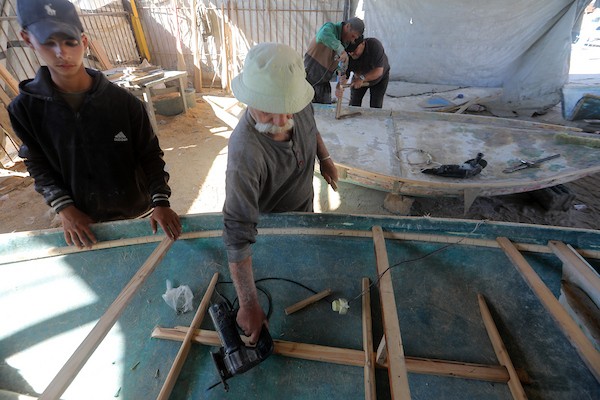 الفلسطيني عطية مقداد وأفراد من عائلته يصلحون قوارب تضررت جراء الغارات الإسرائيلية في ميناء غزة، في 2 يونيو/حزيران 2026. ويستخدمون أخشاباً جرى إنقاذها من المباني المدمّرة لصيانة القوارب وصنع المجاديف.تصوير: بلال أسامة