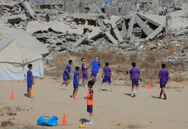 أطفال فلسطينيون نازحون يرتدون الزي الرياضي يشاركون في حصة تدريبية لكرة القدم مع مدربهم في ساحة صغيرة وسط أنقاض المنازل المدمرة في حي الشيخ رضوان بمدينة غزة، 16 حزيران/يونيو 2026. تصوير: بلال أسامة