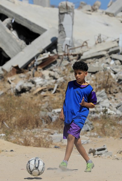 أطفال فلسطينيون نازحون يرتدون الزي الرياضي يشاركون في حصة تدريبية لكرة القدم مع مدربهم في ساحة صغيرة وسط أنقاض المنازل المدمرة في حي الشيخ رضوان بمدينة غزة، 16 حزيران/يونيو 2026. تصوير: بلال أسامة
