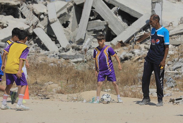 أطفال فلسطينيون نازحون يرتدون الزي الرياضي يشاركون في حصة تدريبية لكرة القدم مع مدربهم في ساحة صغيرة وسط أنقاض المنازل المدمرة في حي الشيخ رضوان بمدينة غزة، 16 حزيران/يونيو 2026. تصوير: بلال أسامة