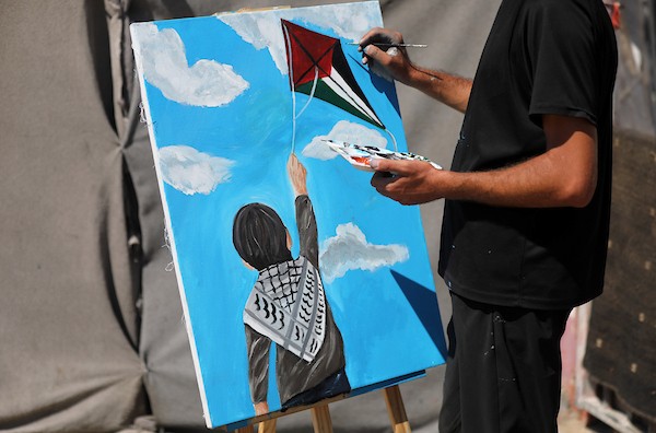الفنان التشكيلي أحمد الحلبي يرسم طفلًا يمسك بخيط طائرة ورقية تحمل ألوان العلم الفلسطيني، أمام خيمة للنازحين في مدينة غزة،  25 أغسطس/آب 2026.  آخر.( تصوير: بلال أسامة )