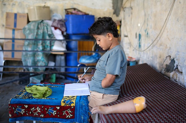 الطفل الفلسطيني كنان أبو جبة، الناجي الوحيد من عائلته بعد استهداف منزلهم في حي الشجاعية، يكتب في دفتره داخل خيمة بمدينة غزة مستخدمًا طرفًا صناعيًا بعد بتر يده اليسرى، 21 سبتمبر/أيلول 2026. تصوير: بلال أسامة.