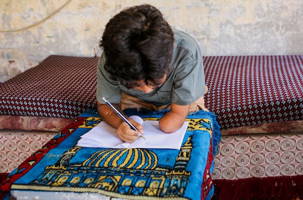 الطفل الفلسطيني كنان أبو جبة، الناجي الوحيد من عائلته بعد استهداف منزلهم في حي الشجاعية، يكتب في دفتره داخل خيمة بمدينة غزة مستخدمًا طرفًا صناعيًا بعد بتر يده اليسرى، 21 سبتمبر/أيلول 2026. تصوير: بلال أسامة.