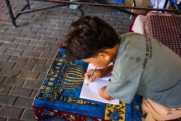 الطفل الفلسطيني كنان أبو جبة، الناجي الوحيد من عائلته بعد استهداف منزلهم في حي الشجاعية، يكتب في دفتره داخل خيمة بمدينة غزة مستخدمًا طرفًا صناعيًا بعد بتر يده اليسرى، 21 سبتمبر/أيلول 2026. تصوير: بلال أسامة.
